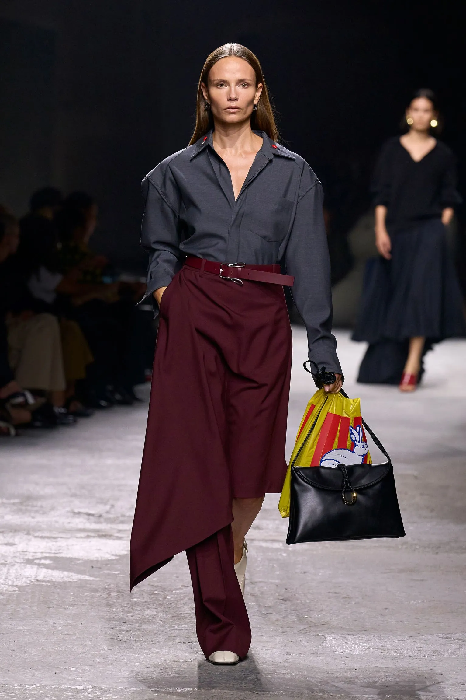 fashion model on runway wearing a gray shirt and maroon pants with bags