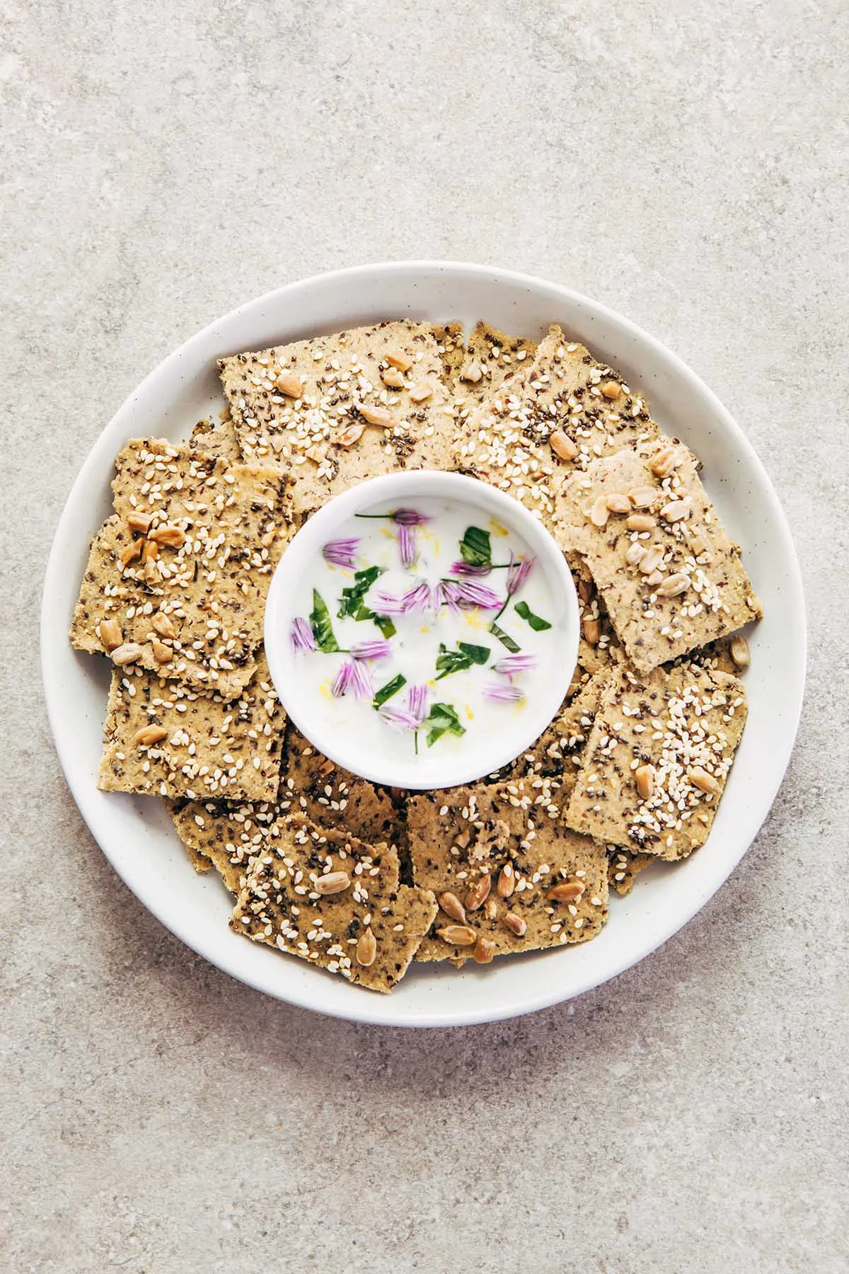 A plate of multi-seed oat flour crackers on a small white plate with a bowl of lemon herb yogurt dip in the middle of the plate.
