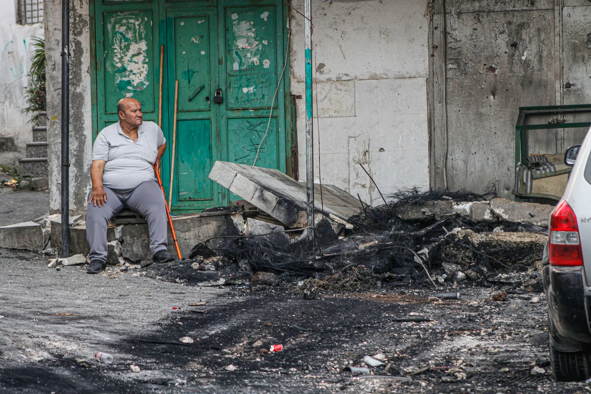 A man sits next to a green door, with remnants of a damaged building next to him.