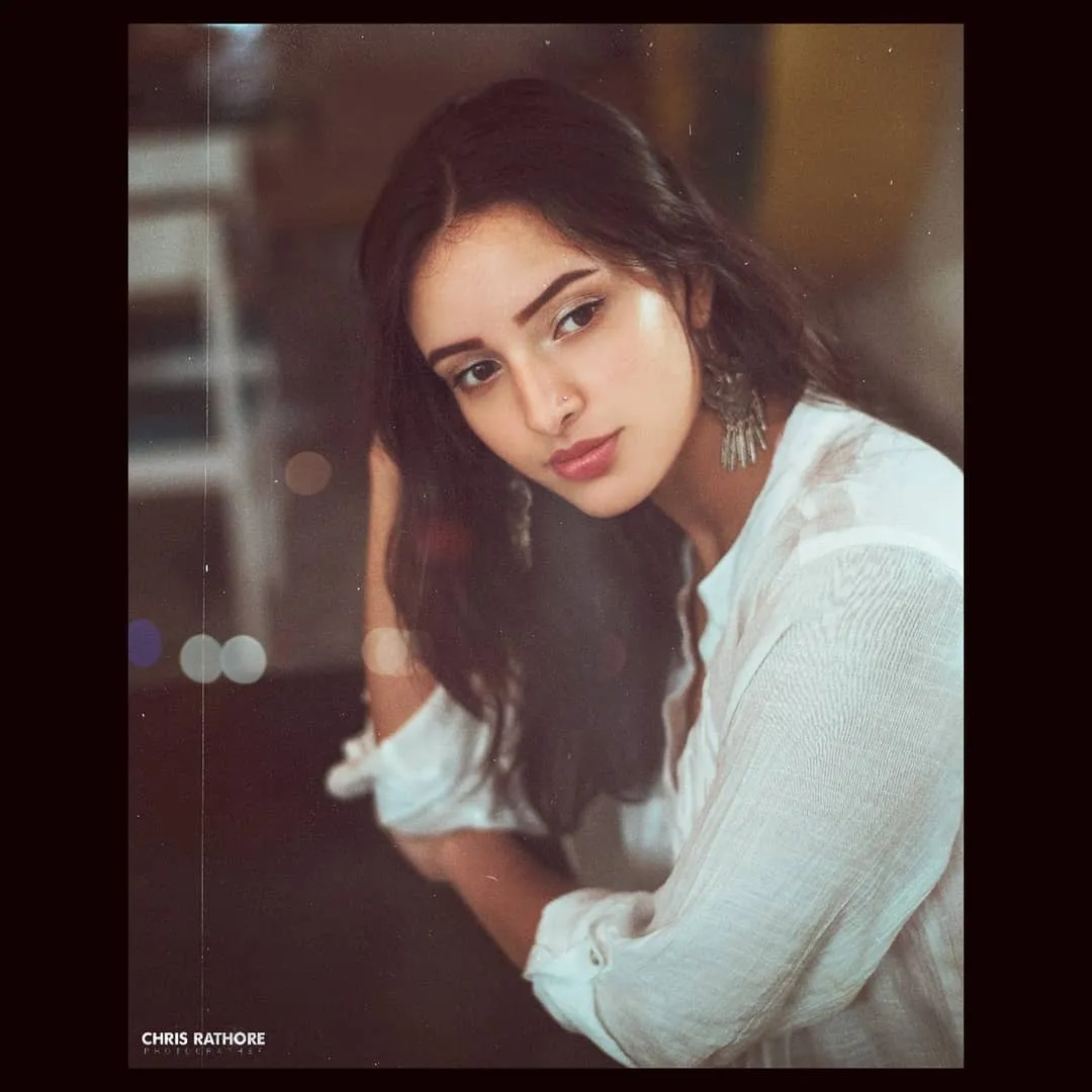 Tripti Dimri in a white top, with light makeup and large earrings. Her head rests on her hand, giving a calm and thoughtful look. The soft lighting adds a warm feeling, and the blurry background makes the scene look ar.jpg  