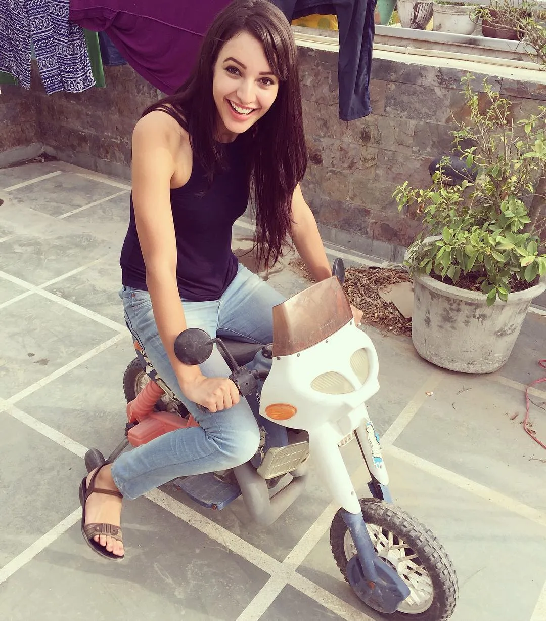 Tripti Dimri is sitting on a small toy motorbike, smiling brightly while enjoying a playful moment. The setting seems to be an outdoor area with plants and hanging clothes in the background, adding a casual and fun vib.jpg  