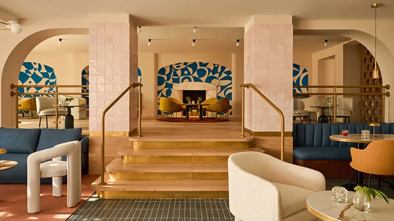 A stylish hotel lounge with a blend of modern and vintage design elements. The space features warm tones, soft lighting, and a mix of seating, including plush chairs and curved sofas. A set of steps with brass accents leads to an elevated area with a fireplace framed by bold blue geometric artwork. Arched openings and decorative columns covered in soft pink tiles add to the aesthetic, while tables with drinks and plants contribute to the cozy ambiance