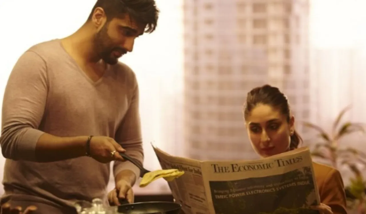 A man and a woman are cooking together in a kitchen, with the woman reading the newspaper and the man looking at her with a smile.