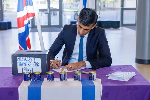 Rishi Sunak writing a card at a table draped in an Israeli flag, next to a box marked 'messages and prayers for Israel'.